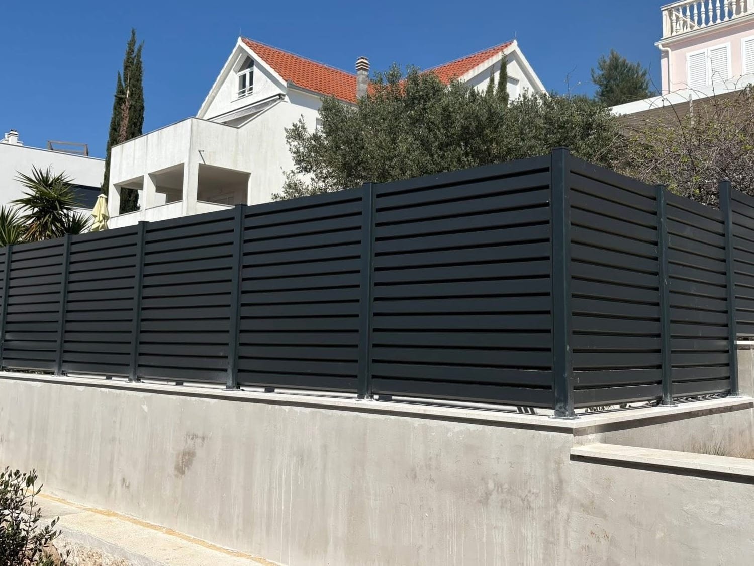Antracitna aluminijska ograda s ravnim horizontalnim lamelama postavljena na betonski zid ispred dalmatinskih kuća.