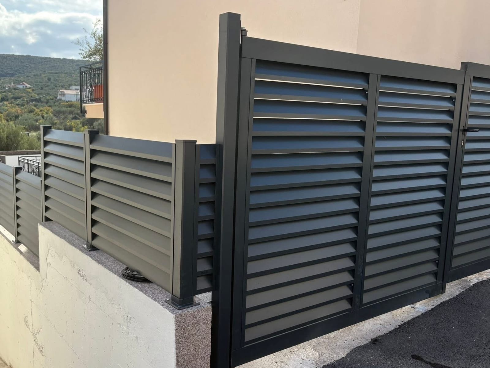 Antracitna aluminijska ograda s kosim lamelama i dvokrilna vrata iste izvedbe uz ulazni prilaz obiteljske kuće.