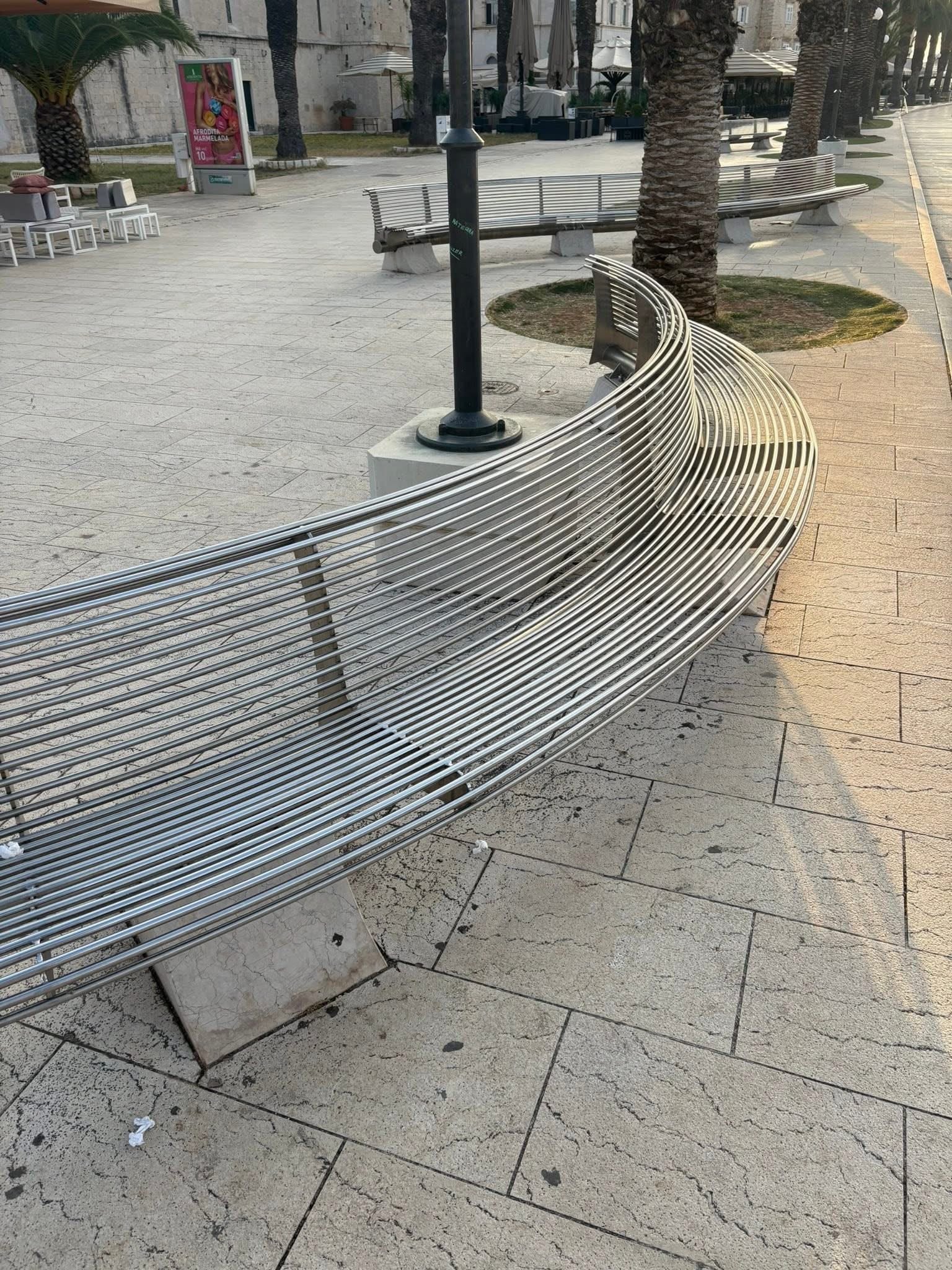 Inox klupa na rivi uz palme i povijesne zgrade u pozadini.