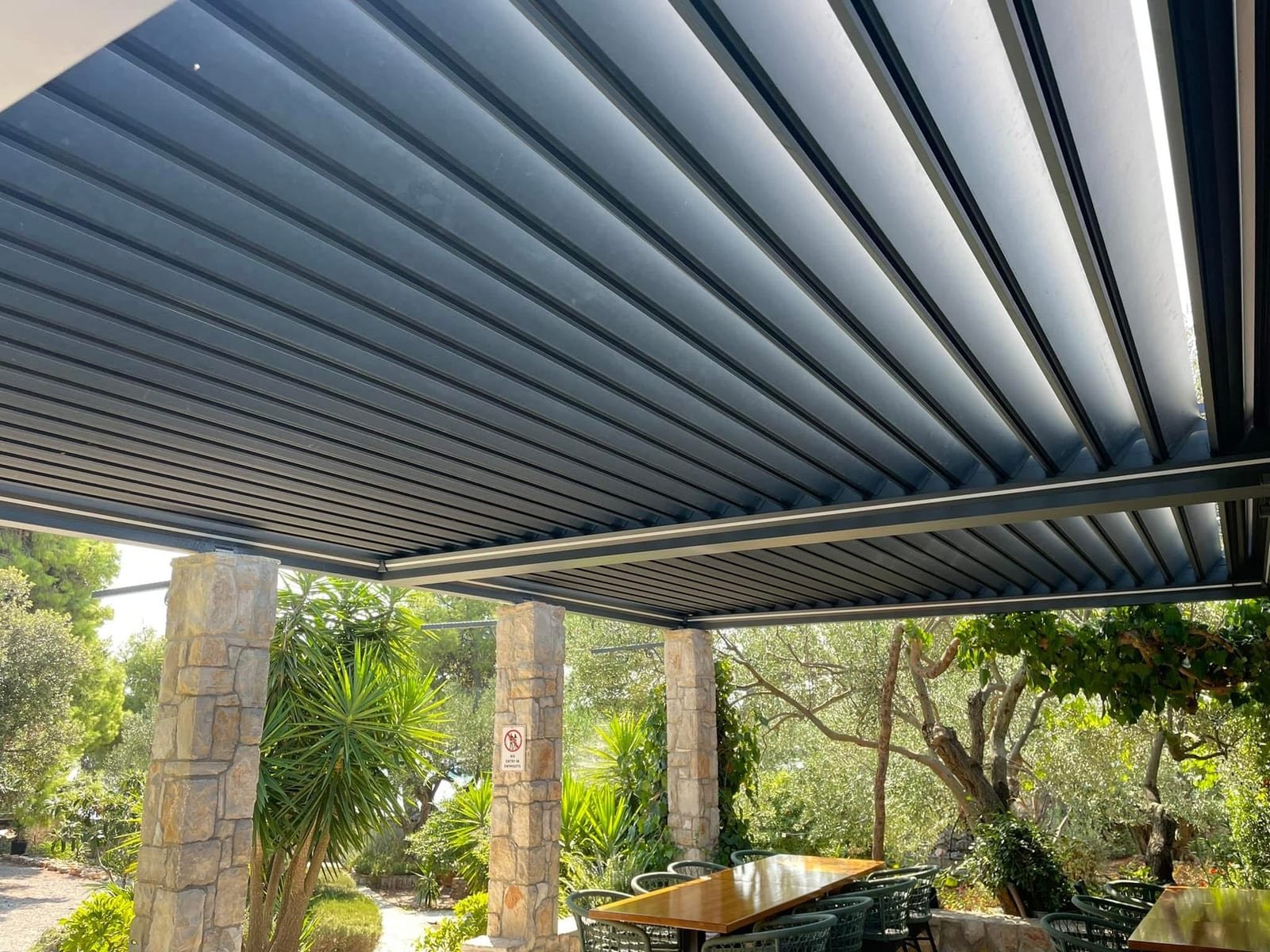 Lamele antracitne aluminijske pergole snimljene odozdo, s kamenim stupovima i vrtom u pozadini.