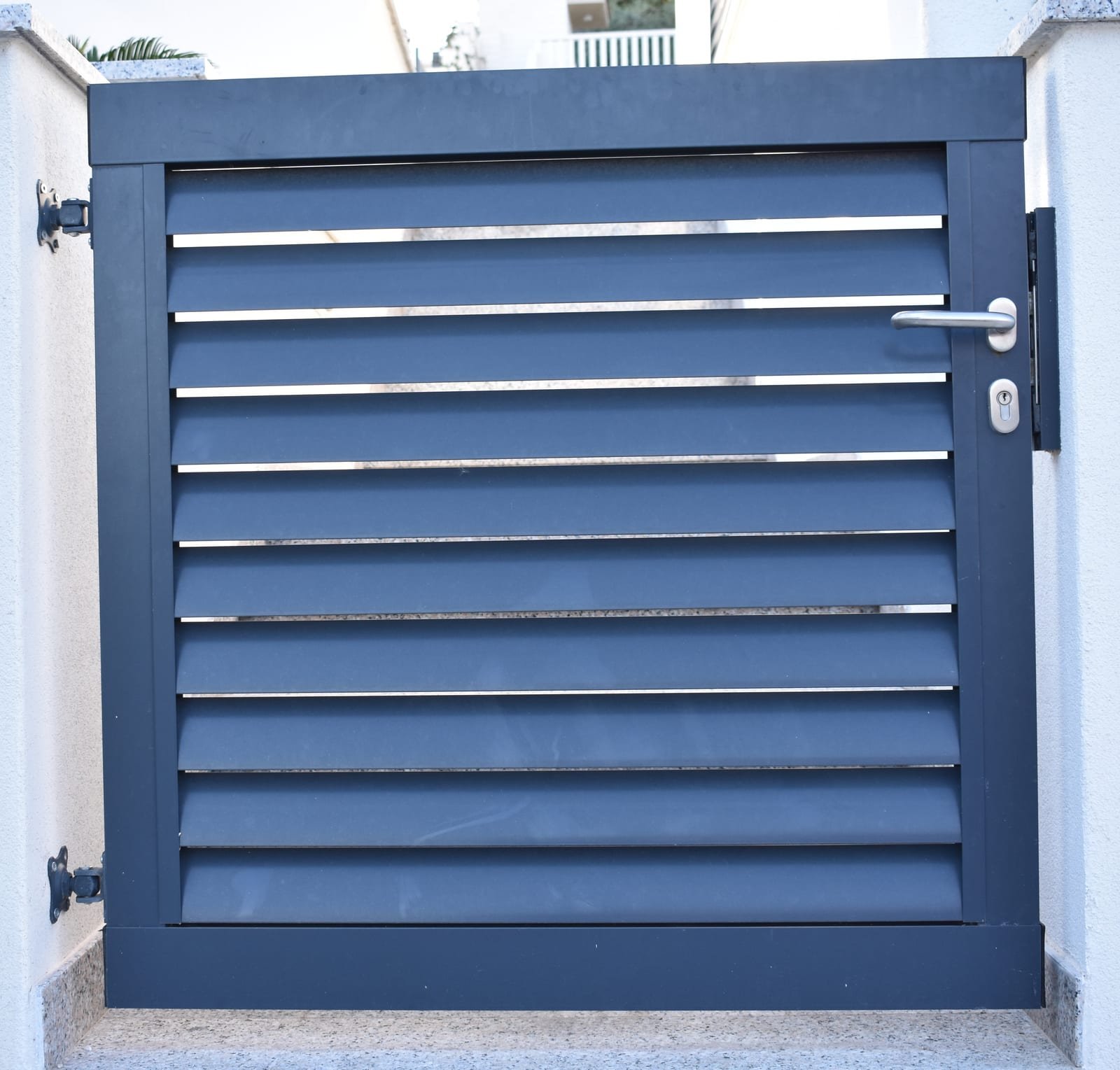 Jednokrilna aluminijska vrata s kosim lamelama u antracit boji ugrađena između bijelih zidanih stupova, s inox kvakom i cilindar bravom.