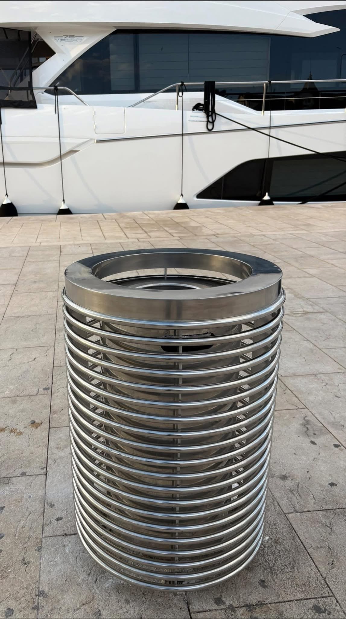Inox koš za otpad s polukružnim obručima na popločanoj rivi ispred velike bijele jahte.