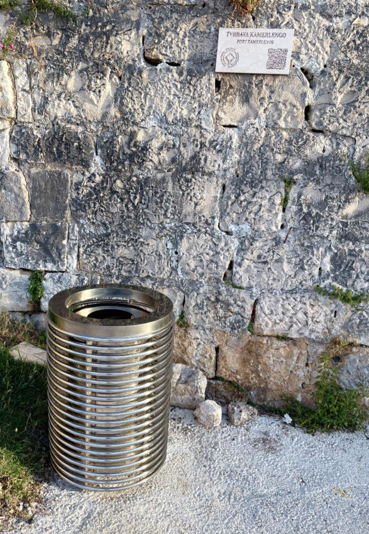 Inox koš za otpad postavljen na šljunku uz grubi kameni zid tvrđave Kamerlengo.