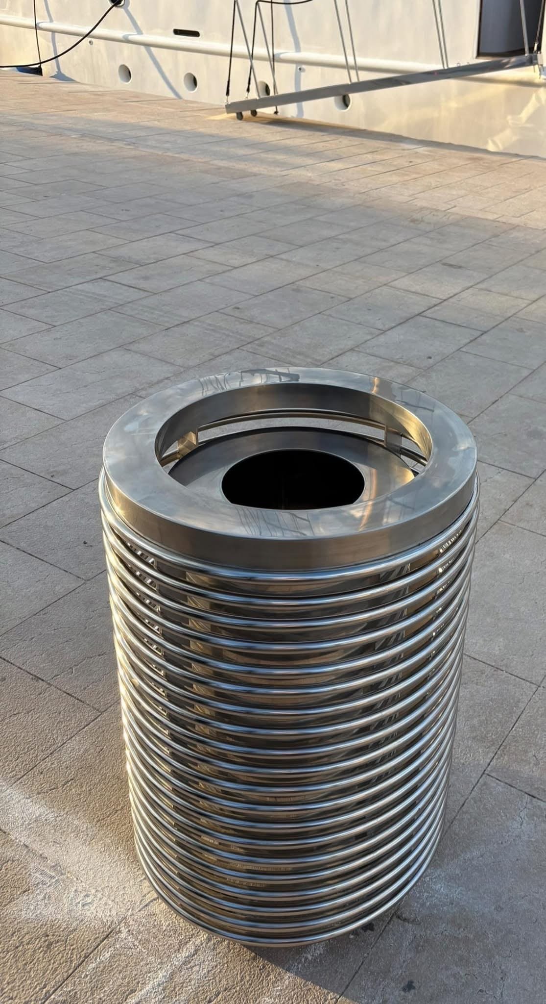 Inox koš za otpad na popločanoj rivi u toplom večernjem svjetlu, s pramcem privezane jedrilice u pozadini.