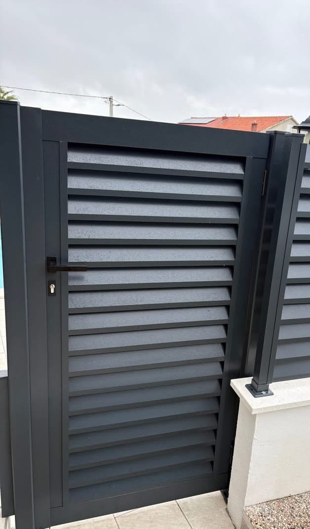 Jednokrilna vrata s kosim aluminijskim lamelama u antracit boji, s kvakom i cilindar bravom, uz ogradni panel iste izvedbe.