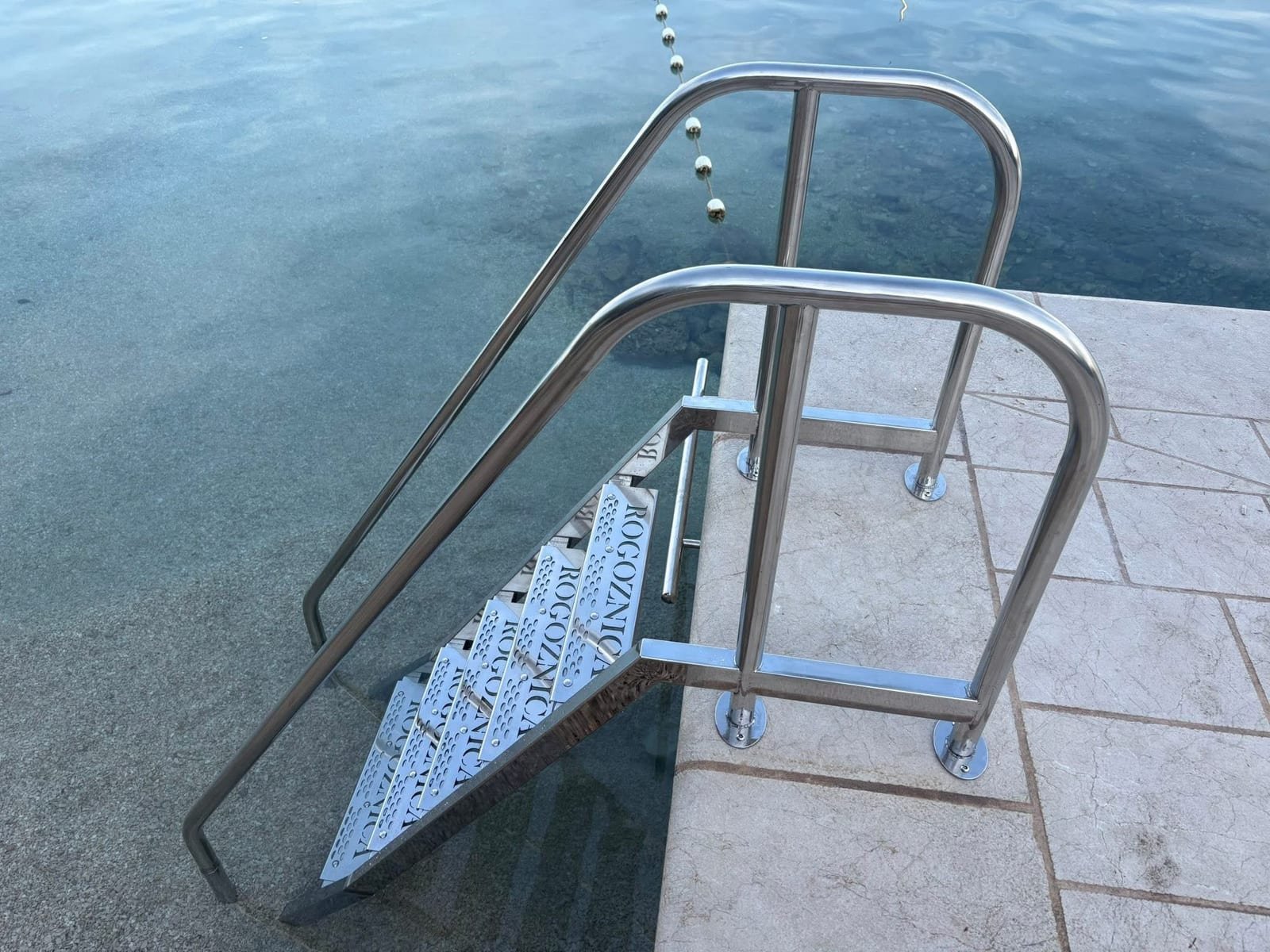 Inox kupališne stepenice s perforiranim gazištima i dvostrukim rukohvatima na rubu betonske rive.