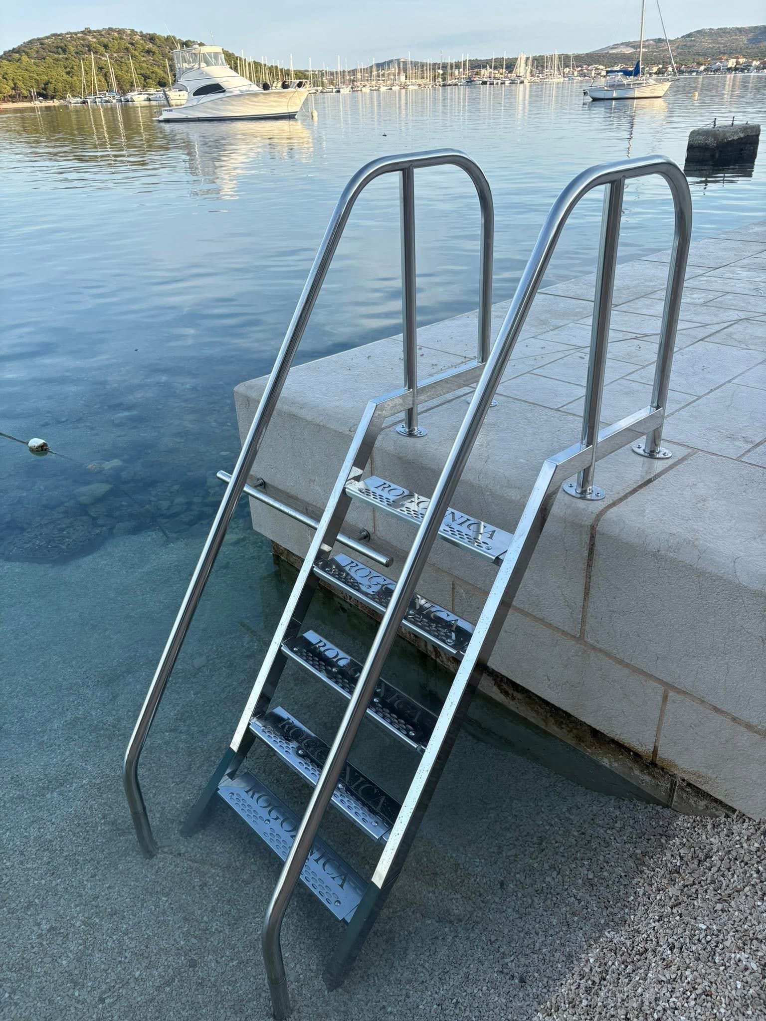Inox ljestve za ulaz u more postavljene na rivi, s pogledom na marinu i usidrene brodove.