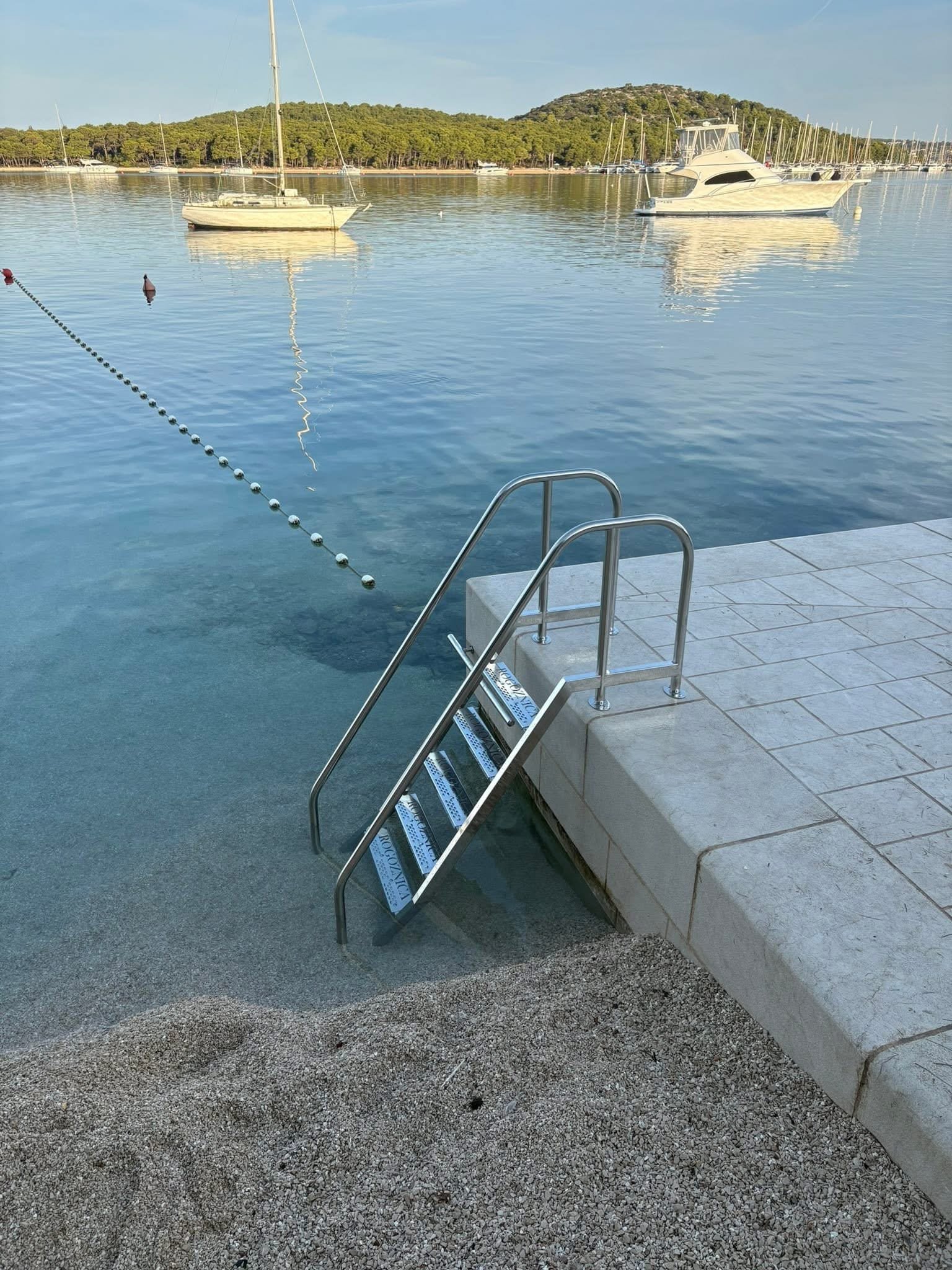 Mirna uvala s usidrenim jedrilicama i inox kupališnim stepenicama na betonskoj rivi.