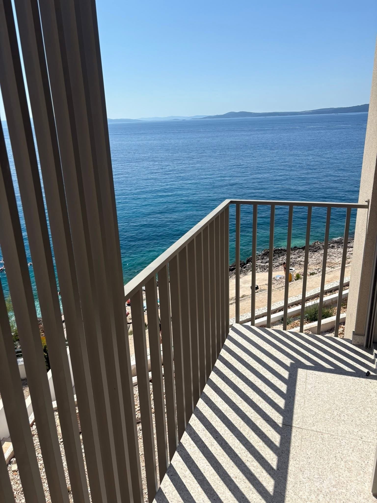 Pogled s balkona kroz aluminijske lamele na more i kamenu plažu.