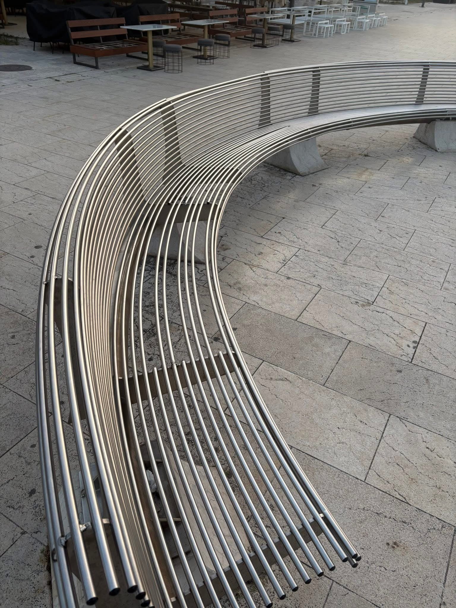 Valovita inox klupa u obliku slova S postavljena na kamenom trgu ispred ugostiteljske terase.