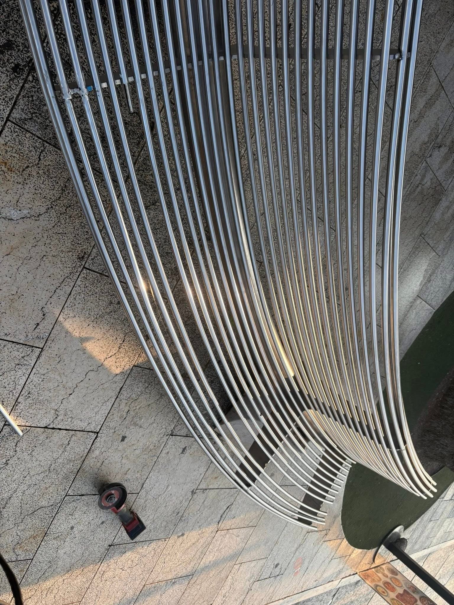Detalj savijenih inox cijevi klupe koje se spuštaju prema tlu, s kutnom brusilicom odloženom na popločenju.