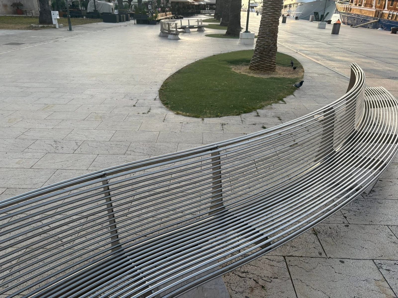 Duga inox klupa prati rub rive, iza nje palma na travnatom otoku i privezani brod.