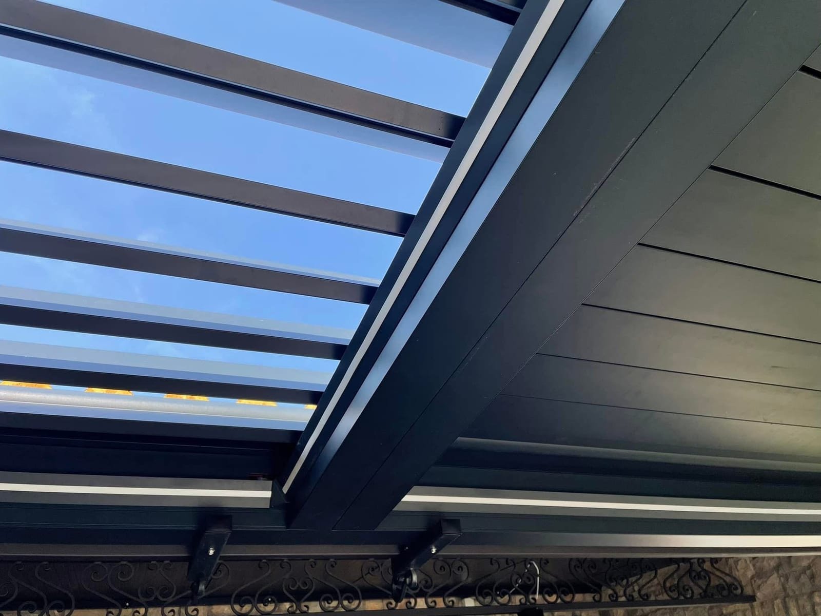 Detalj spoja nosive grede i lamela aluminijske pergole s ugrađenom LED trakom, snimljen prema nebu.