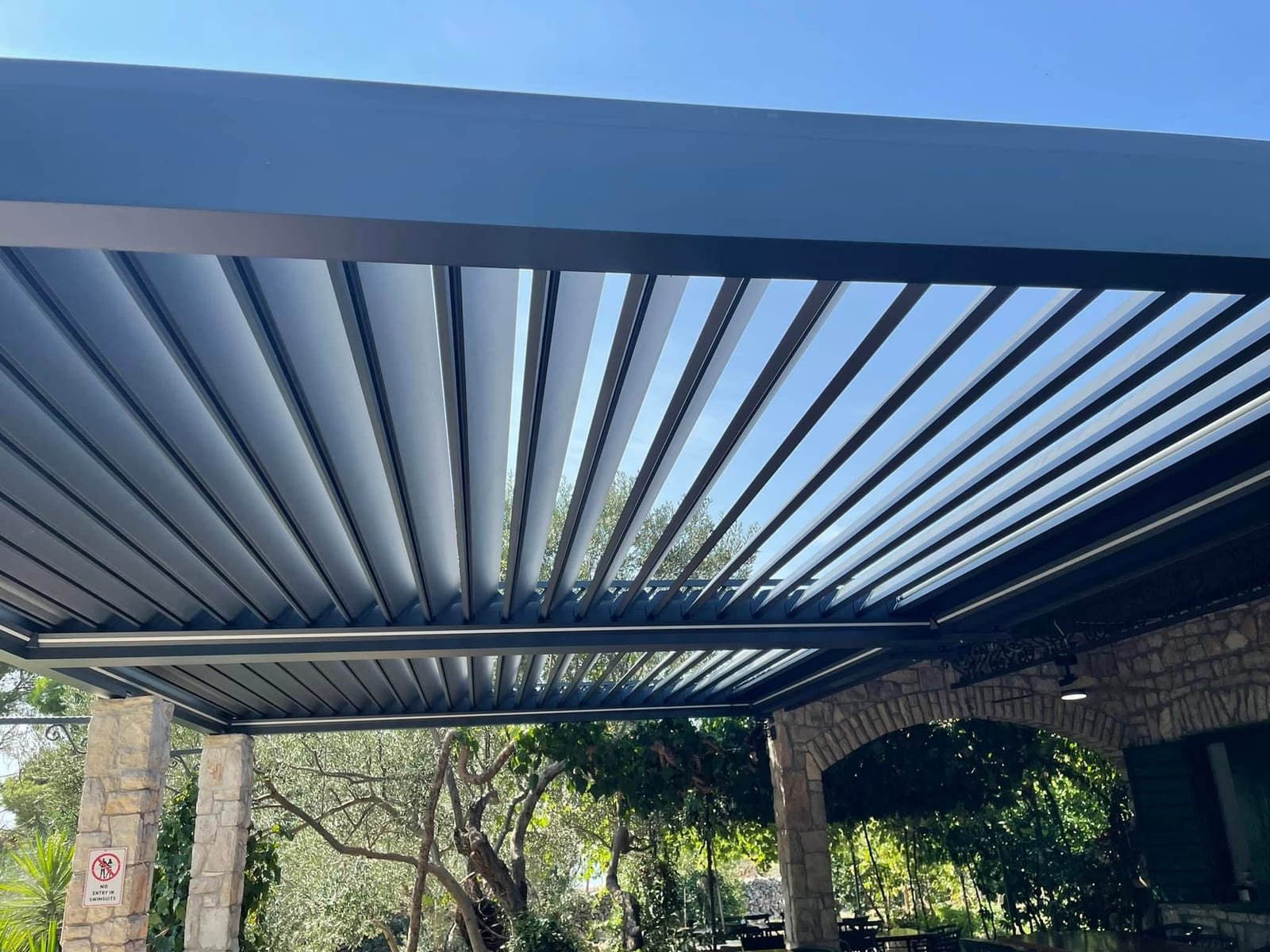 Pogled odozdo na otvorene lamele aluminijske pergole kroz koje se vidi vedro plavo nebo.