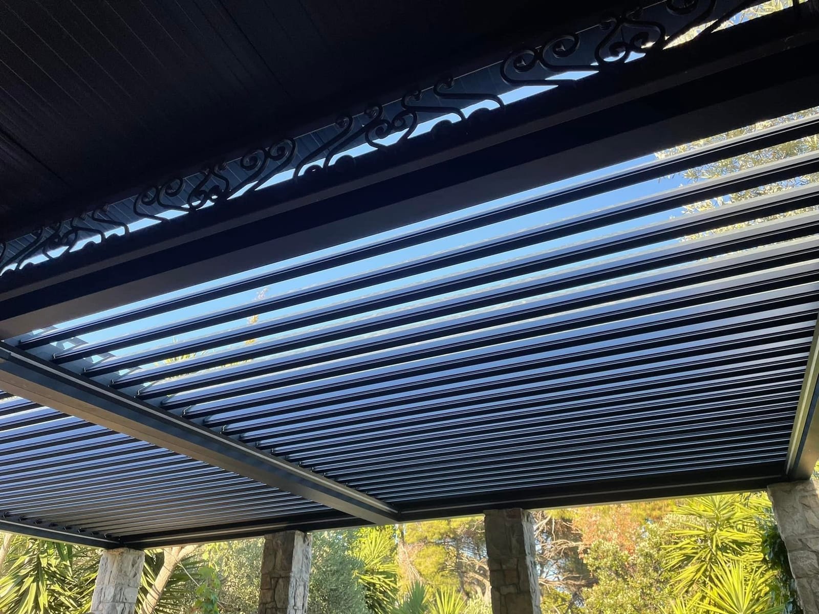 Aluminijska pergola sa zatvorenim lamelama snimljena odozdo, s kamenim stupovima i zelenilom u pozadini.