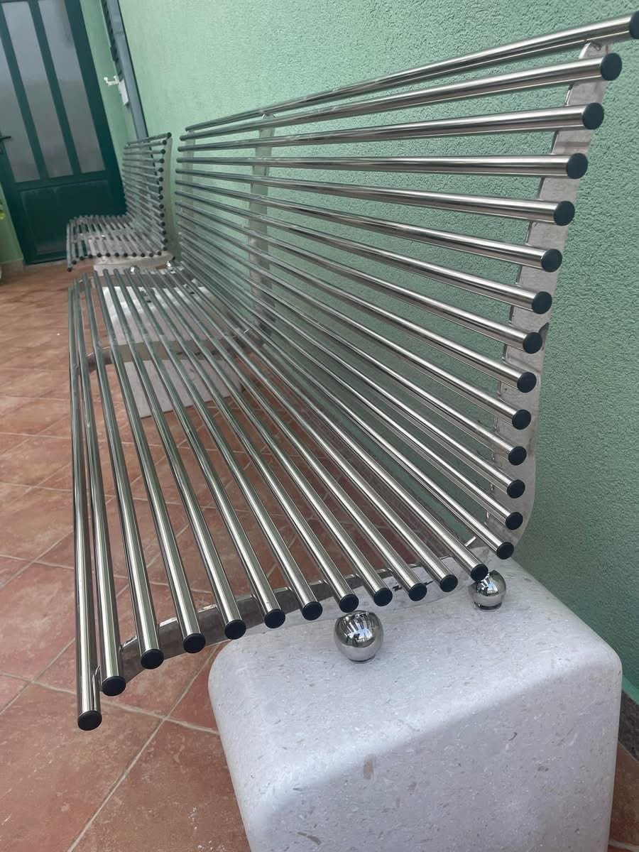 Klupa od inox cijevi s crnim čepovima na kamenim postoljima uz zeleni zid natkrivenog trijema.