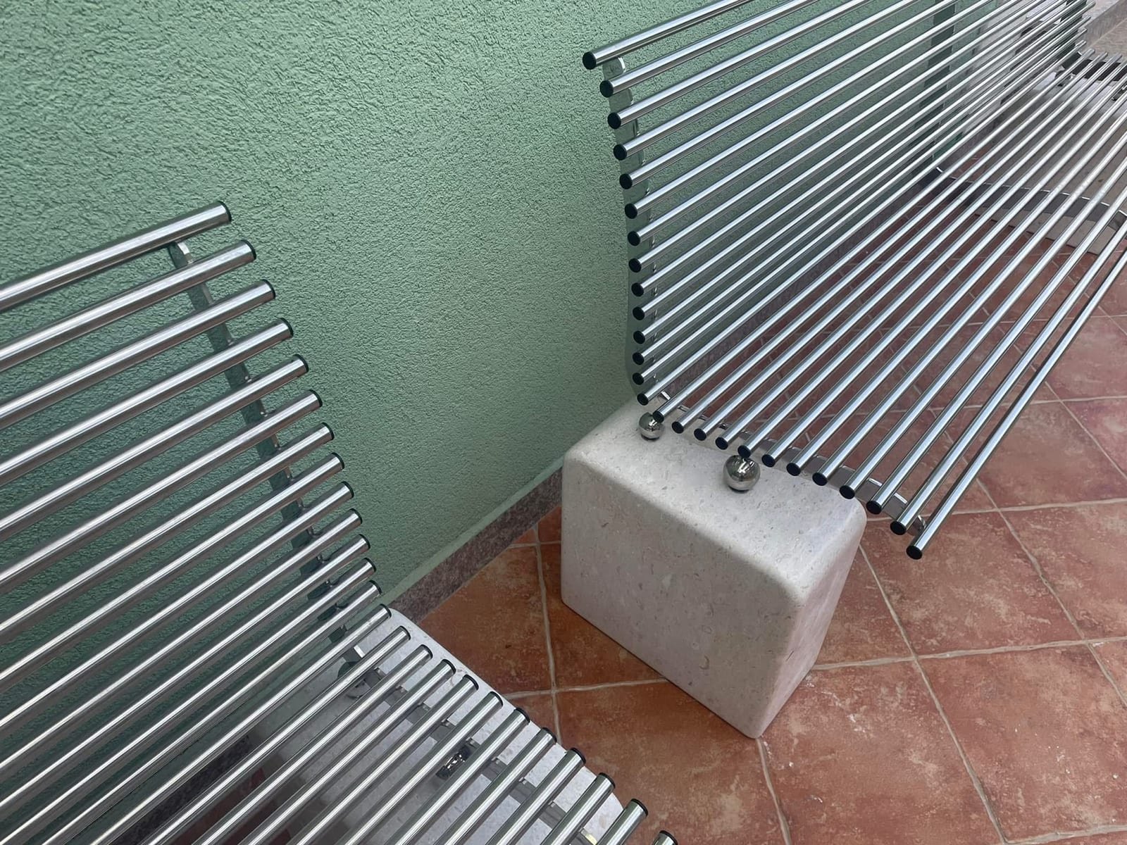 Detalj inox klupe od nehrđajućih cijevi s kamenim postoljem i poliranim kuglastim pričvrsnicima uz zelenu fasadu.