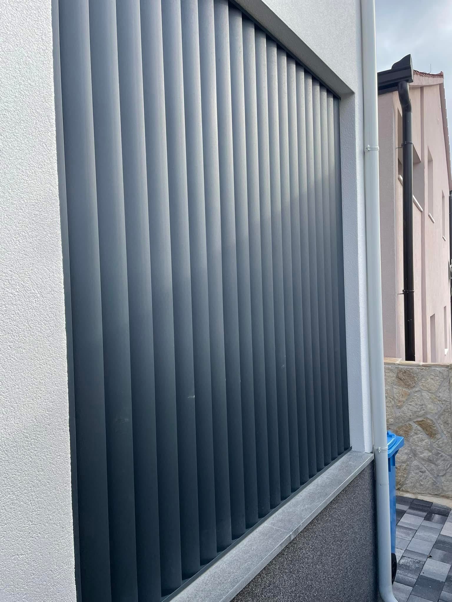 Antracitna ograda od punih aluminijskih lamela postavljena na niski zid uz bijelo pročelje kuće.